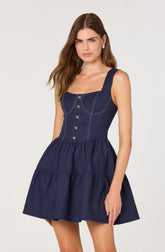 A woman wearing a sleeveless, navy blue dress with wide shoulder straps. The dress has contrast stitching details, a fitted bodice with button-down front, and a gathered, tiered skirt.